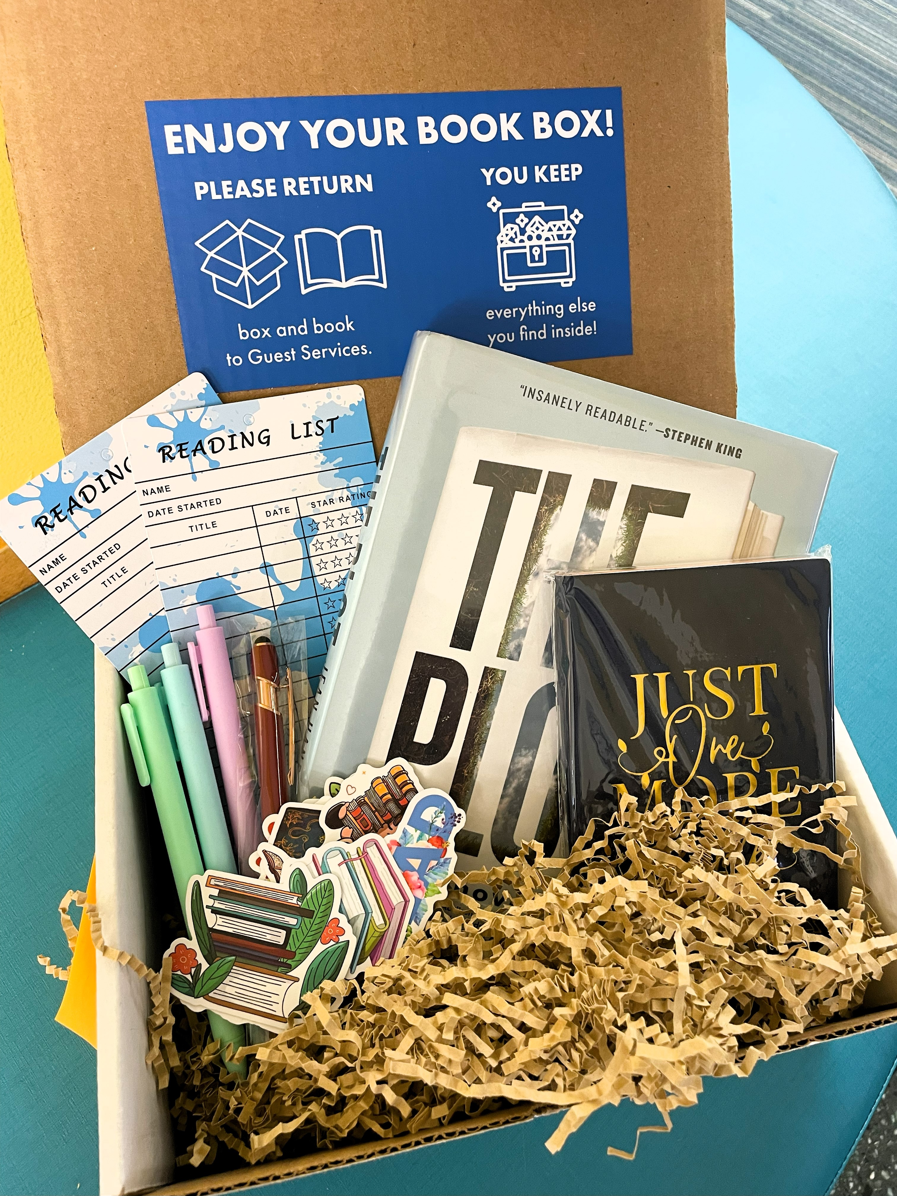 Photo of the inside of a Book Box. Cardboard box with stickers, pens, book marks, a journal, and a book.