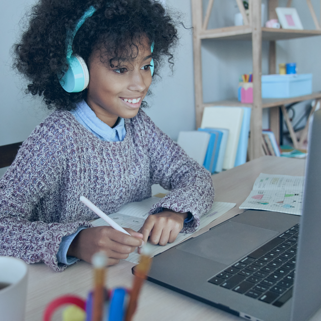 Stock photo. Young adult female presenting with dark skin and textured hair sitting at laptop, listening with headphones, and writing in a journal.