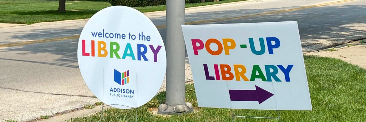 two signs staked in the grass near a curb, one reading "welcome to the library" and the other reading "pop-up library" with an arrow pointing right