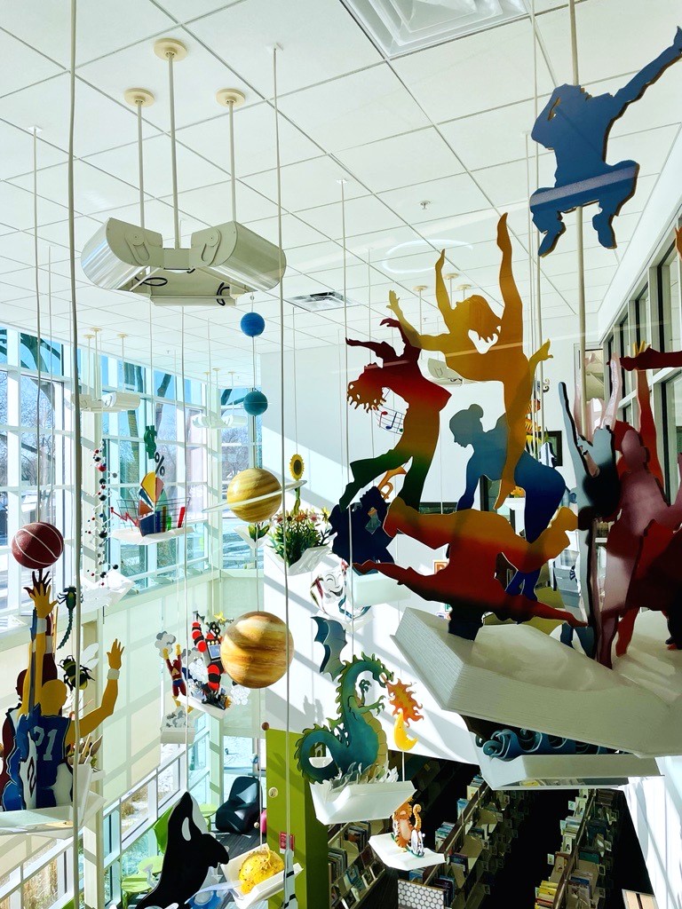 Colorful hanging decorations depicting sports, planets, and mythical creatures are suspended from the ceiling in a bright library space with large windows.