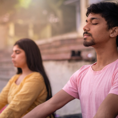 Photo of two adults meditating. One male presenting in a pink shirt. The other female presenting in a yellow shirt. Both have their eyes closed.