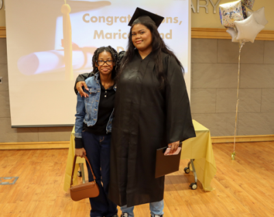 photo of a graduate in a black cap and gown standing with a family member in front of a screen that says "Congratulations!"