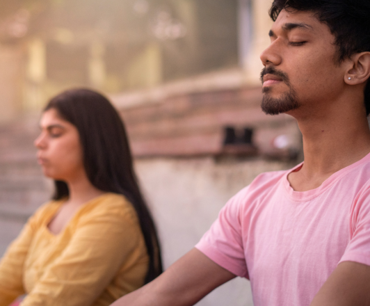 Photo of two adults meditating. One male presenting in a pink shirt. The other female presenting in a yellow shirt. Both have their eyes closed.