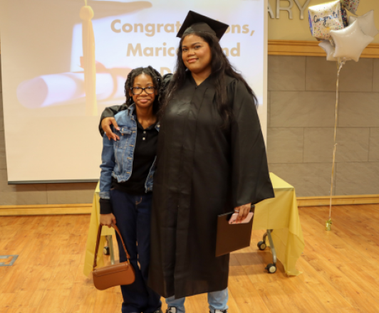 photo of a graduate in a black cap and gown standing with a family member in front of a screen that says "Congratulations!"