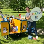 Staff member holding a sign reading "Welcome to the Library" in front of the book bike.