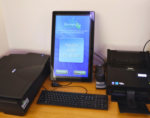 Self-service scanning station with touchscreen monitor displaying 'ScanEZ,' with a prompt to 'Touch Here to Start.' The station includes a large flatbed scanner on the left, a document feeder scanner on the right, a keyboard, and a mouse.