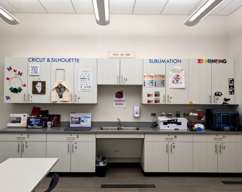 Photo of Creative Studio counter and cabinets. Sitting on counter is 3D printer, sublimation printer, and sewing machine. Cabinets are displaying projects like bags and 3D prints.