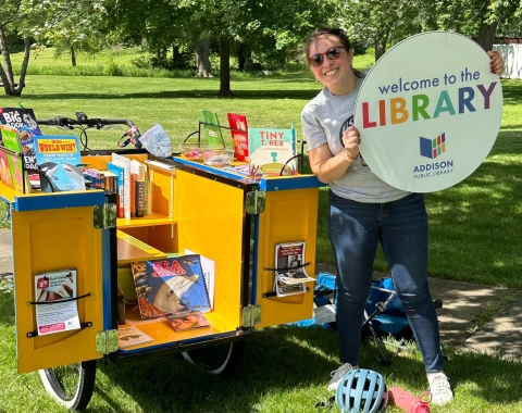 Staff member holding a sign reading "Welcome to the Library" in front of the book bike.