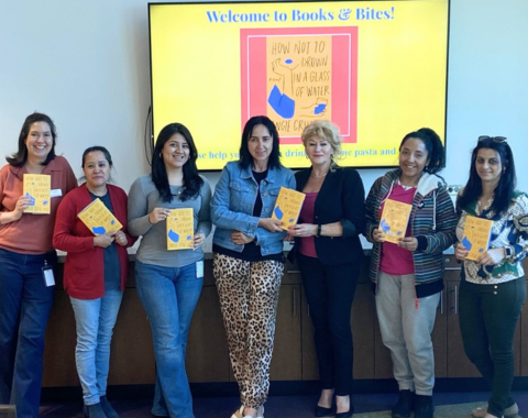 seven people posing for photo in front of tv monitor that displays "Welcome to Books & Bites" and they are all holding copies of the book "How Not to Drown in a Glass of Water" by Angie Cruz