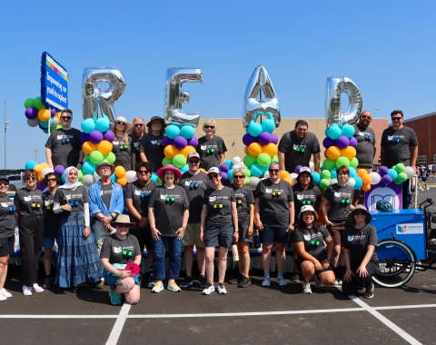 Large group of people standing on and around parade float with large silver balloons spelling "READ"